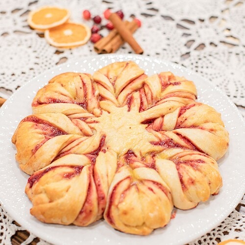 A sourdough cranberry orange star bread on a white plate showing the golden twisted star shape with deep red cranberry filling, styled on a lace doily with dried orange slices, fresh cranberries, and cinnamon sticks in a cottage kitchen.