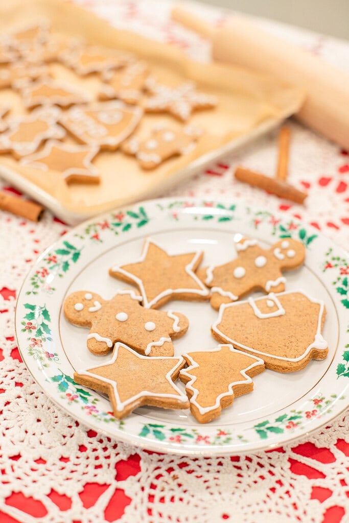 A holly-rimmed vintage china plate with sourdough gingerbread cookies in gingerbread man, star, Christmas tree, and Santa shapes decorated with white royal icing, with a parchment-lined baking sheet of more cookies, a rolling pin, and cinnamon sticks in the background.