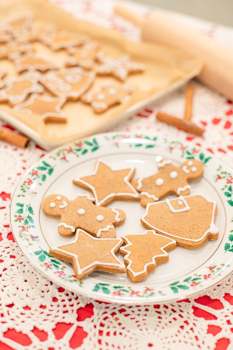 A holly-rimmed vintage china plate with sourdough gingerbread cookies in gingerbread man, star, Christmas tree, and Santa shapes decorated with white royal icing, with a parchment-lined baking sheet of more cookies, a rolling pin, and cinnamon sticks in the background.