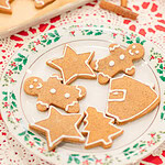 A holly-rimmed vintage china plate of sourdough gingerbread cookies decorated with white royal icing in star, gingerbread man, Christmas tree, and Santa shapes, with a baking sheet of more decorated cookies and a rolling pin softly blurred behind it on a red lace doily.
