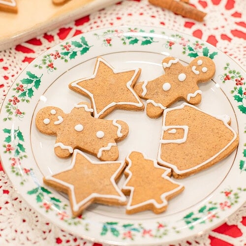 A holly-rimmed vintage china plate of sourdough gingerbread cookies decorated with white royal icing in star, gingerbread man, Christmas tree, and Santa shapes, with a baking sheet of more decorated cookies and a rolling pin softly blurred behind it on a red lace doily.
