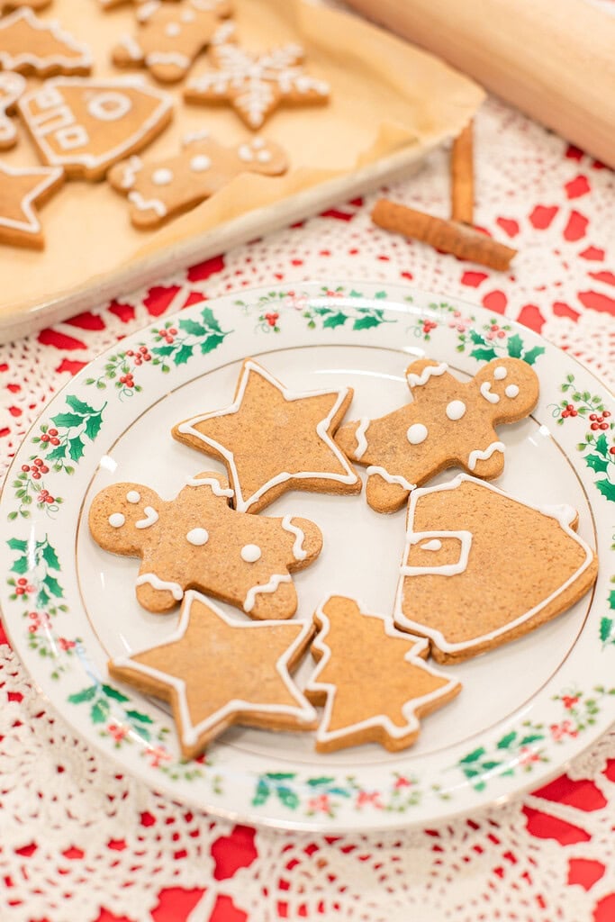 A holly-rimmed vintage china plate of sourdough gingerbread cookies decorated with white royal icing in star, gingerbread man, Christmas tree, and Santa shapes, with a baking sheet of more decorated cookies and a rolling pin softly blurred behind it on a red lace doily.