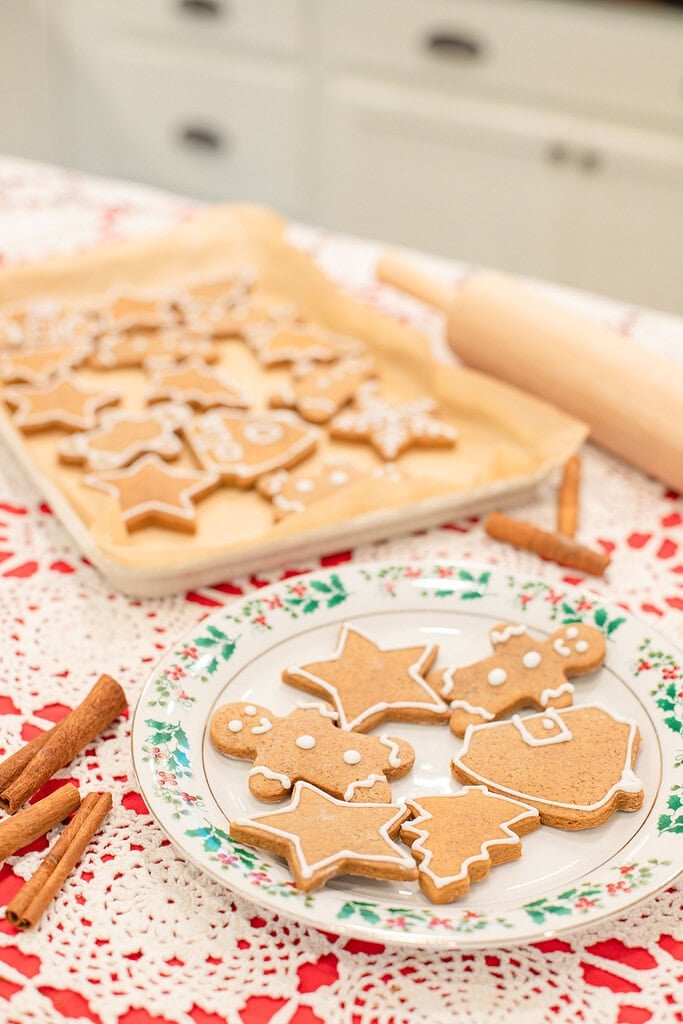 A holly-rimmed vintage china plate of iced sourdough gingerbread cookies in the foreground, with a full parchment-lined baking sheet of decorated Christmas-shaped cookies, cinnamon sticks, and a rolling pin in a cottage kitchen.