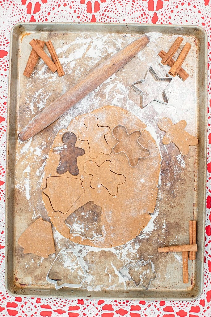 Overhead view of rolled sourdough gingerbread cookie dough on a flour-dusted baking sheet with gingerbread man, Christmas tree, and star shapes cut out, surrounded by metal cookie cutters and cinnamon sticks on a red lace doily.
