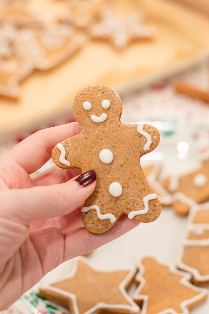 A hand holding a sourdough gingerbread man cookie decorated with white royal icing dots and squiggle details, with a baking sheet of more decorated gingerbread cookies and a holly-trimmed plate softly blurred in the background.