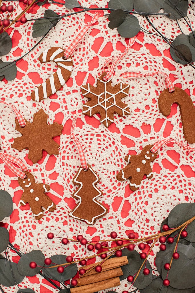 Overhead view of cinnamon applesauce ornaments in snowflake, gingerbread man, Christmas tree, candy cane, and star shapes decorated with white icing and tied with red gingham ribbons on a red and white lace doily with red berry branches, eucalyptus, and cinnamon sticks.