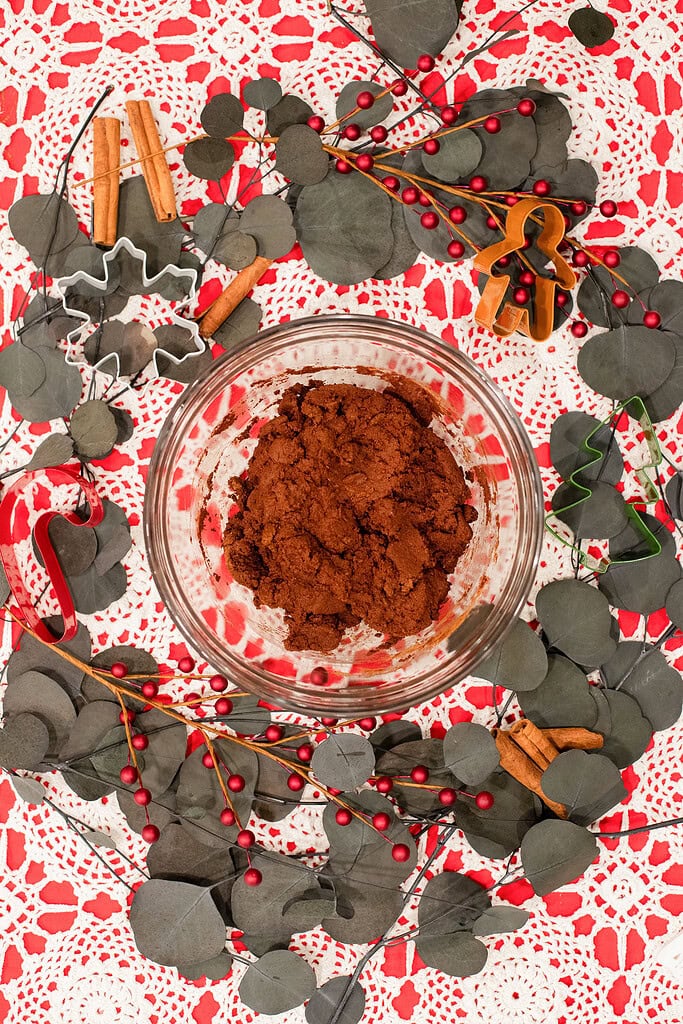 Overhead view of cinnamon applesauce dough mixed in a glass bowl, surrounded by Christmas cookie cutters, cinnamon sticks, red berry branches, and eucalyptus stems on a red and white lace doily.