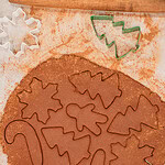 Overhead view of cinnamon applesauce ornament dough rolled out on parchment with Christmas tree, candy cane, and other holiday shapes cut out, surrounded by colorful cookie cutters and a rolling pin.