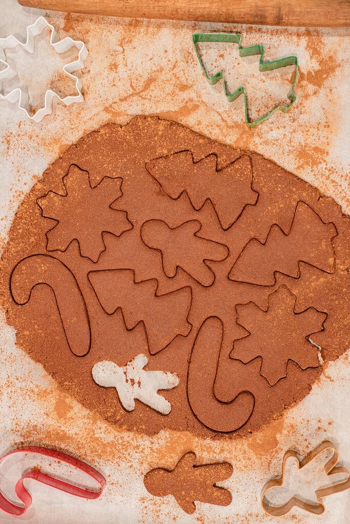 Overhead view of cinnamon applesauce ornament dough rolled out on parchment with Christmas tree, candy cane, and other holiday shapes cut out, surrounded by colorful cookie cutters and a rolling pin.