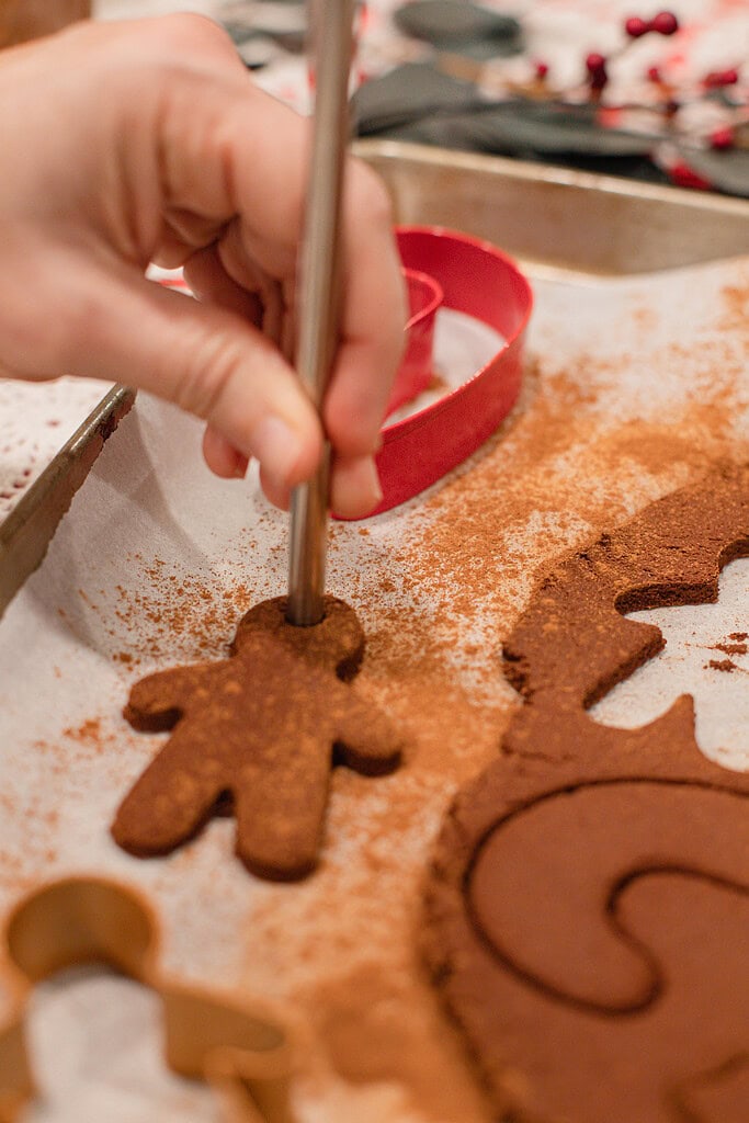A hand using a metal skewer to punch a hanging hole through a freshly cut cinnamon applesauce gingerbread man ornament on a parchment-lined baking sheet, with other cut shapes and a red candy cane cookie cutter visible nearby.