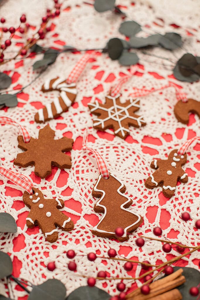 Cinnamon applesauce ornaments in gingerbread man, snowflake, Christmas tree, and candy cane shapes decorated with white icing and tied with red gingham ribbons on a red and white lace doily with red berry branches and eucalyptus.