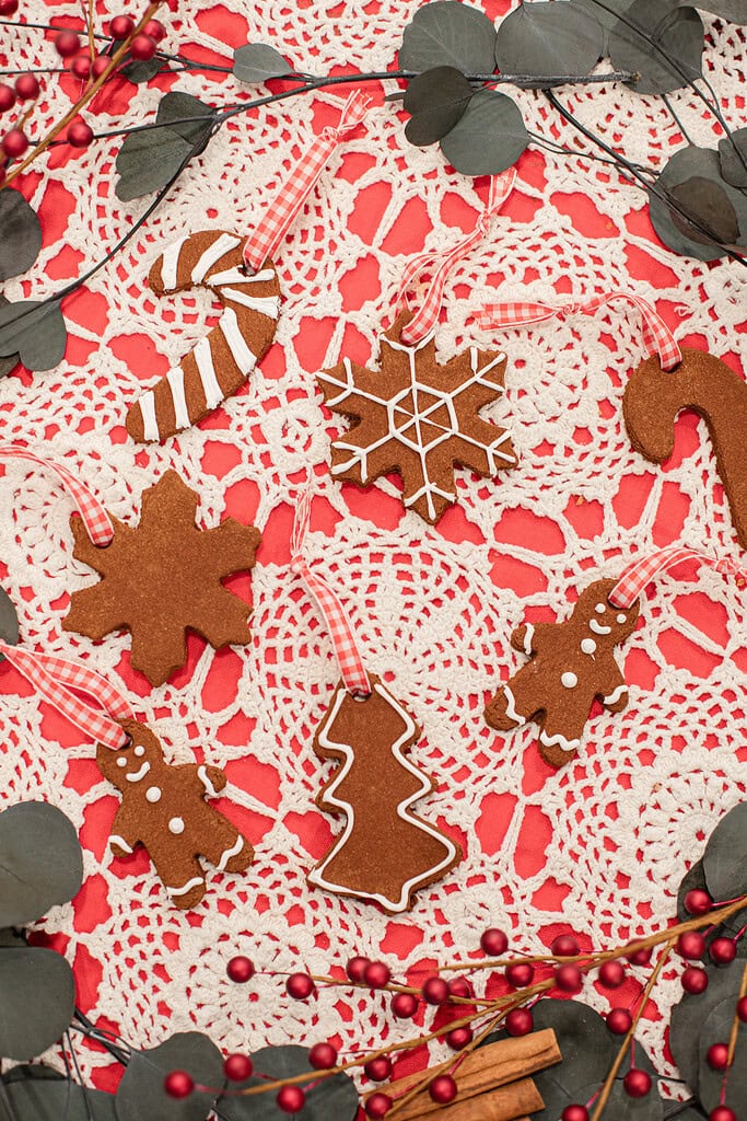 Overhead view of finished cinnamon applesauce ornaments in candy cane, snowflake, gingerbread man, Christmas tree, and candy cane shapes decorated with white icing and hung with red gingham ribbons on a red and white lace doily with red berry branches and eucalyptus.