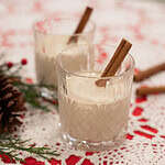 Two glasses of creamy homemade eggnog garnished with cinnamon sticks, placed on a festive holiday table with pine sprigs.
