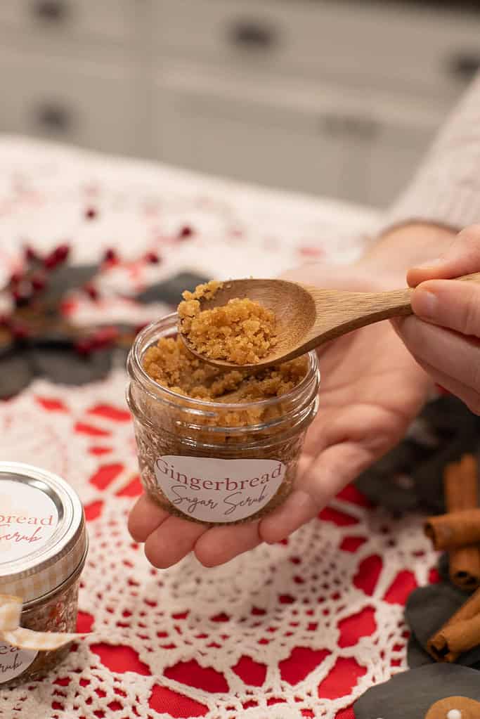 A hand scooping gingerbread-scented sugar scrub into a small jar with holiday labels.