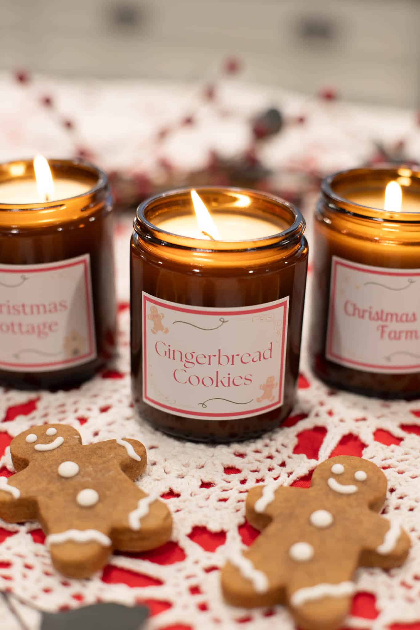 DIY soy wax candles in amber jars labeled "Gingerbread Cookies" and "Christmas Cottage", "Christmas Tree Farm", glowing warmly on a festive table.
