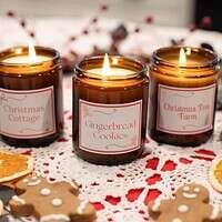 A trio of DIY soy wax candles labeled "Christmas Cottage," "Winter Chai Tea," and "Gingerbread Cookies," styled with festive decor including cinnamon sticks and dried orange slices.