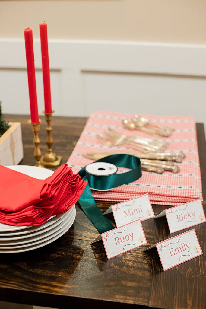 Flat lay of Christmas table setting supplies including stacked white plates, folded red linen napkins, green satin ribbon spool, silver flatware, red gingham placemats, and handwritten place cards reading Mimi, Ruby, Ricky, and Emily