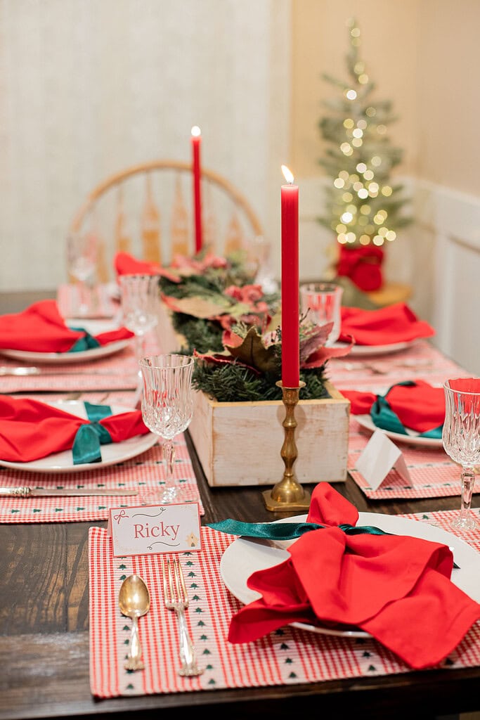 Christmas dinner table set with red gingham placemats, red cloth napkins with green ribbon bows, crystal goblets, gold flatware, a white wood greenery centerpiece with poinsettias and lit red taper candles, and a glowing mini tree in the corner
