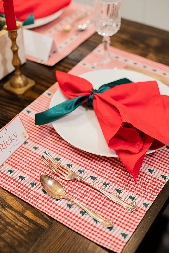 Close-up of a Christmas place setting with a red linen napkin tied in a green satin bow on a white plate, red gingham Christmas tree placemat, gold spoon and fork, and a place card reading Ricky