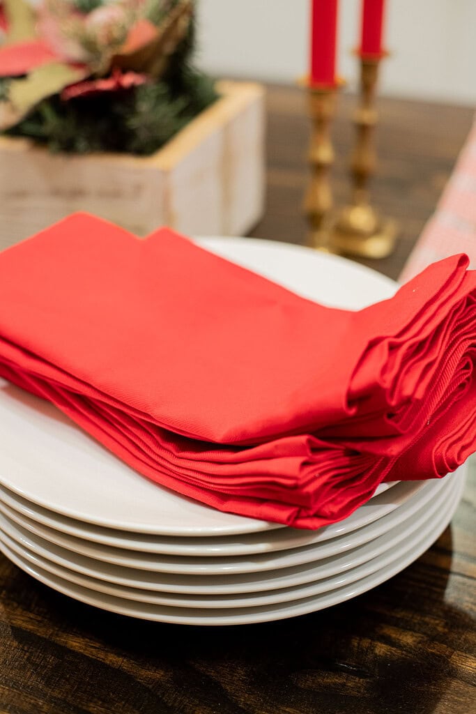 Stack of white dinner plates with folded red linen napkins on a dark wood table, with a white wood greenery centerpiece box and brass taper candle holders blurred in the background