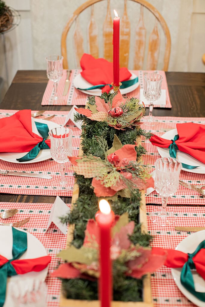 Overhead view of a Christmas table with red gingham placemats, red napkins tied with green ribbon, crystal glasses, silver flatware, and a long white wood centerpiece box filled with evergreen sprigs and poinsettias with two lit red taper candles