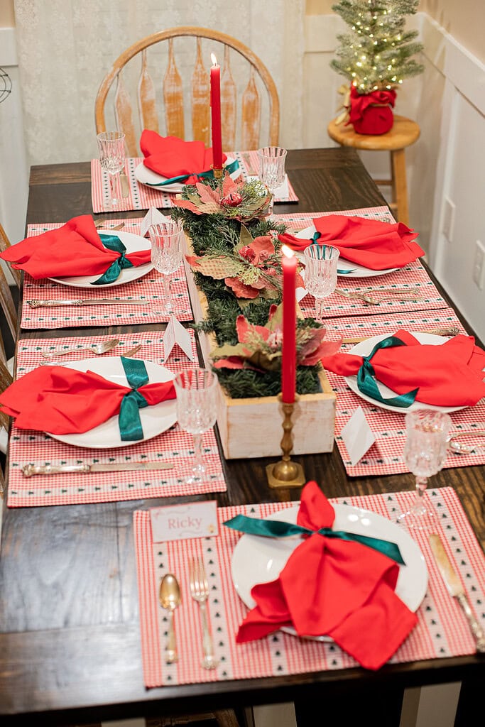 Full overhead view of a Christmas dinner table set for six with red gingham placemats, red linen napkins with green ribbon bows, crystal goblets, gold flatware, place cards, a long greenery and poinsettia centerpiece, and a lit mini Christmas tree on a wooden stool in the background