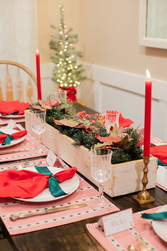 Christmas table detail showing a white distressed wood centerpiece box filled with evergreen sprigs and poinsettias flanked by lit red taper candles in brass holders, red gingham placemats, red napkins, and a place card reading Emily, with a glowing mini tree in the background