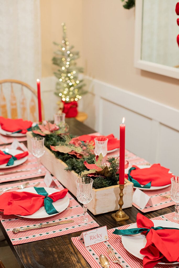 Side view of a Christmas dining table with red gingham placemats, red cloth napkins tied with green satin ribbon, crystal glasses, gold flatware, a white wood greenery centerpiece with poinsettias and a lit red taper candle, place card reading Ricky, and a glowing mini tree in the background