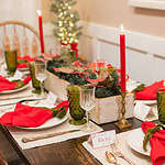 A cozy Christmas table setting featuring red napkins tied with green ribbon on white plates, green glass tumblers, crystal glasses, and gold utensils. A wooden box centerpiece is adorned with pine greenery, red poinsettia accents, and candles. A small Christmas tree with lights is visible in the background.