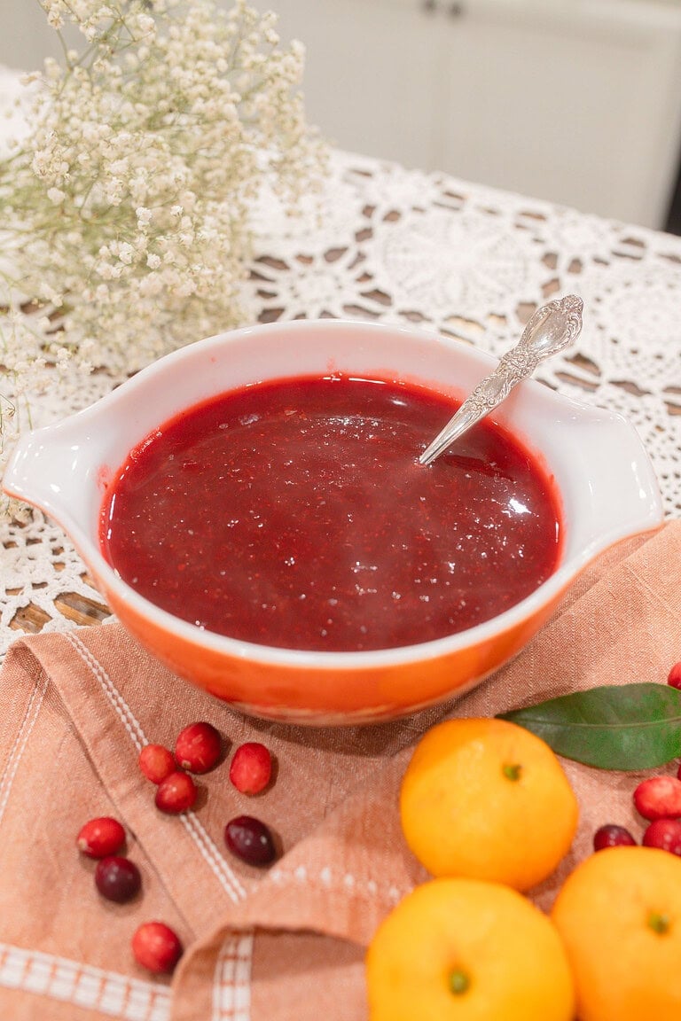 A vintage orange and white Pyrex dish filled with homemade whole berry cranberry orange sauce and a silver spoon, styled on a dusty rose linen with fresh cranberries, whole mandarin oranges, a bay leaf, and dried baby's breath on a lace doily.