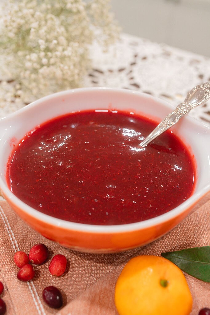 A close-up of a vintage orange and white Pyrex dish filled with glossy homemade cranberry orange sauce and a silver spoon, styled on a dusty rose linen with fresh cranberries, a mandarin orange, and a bay leaf.