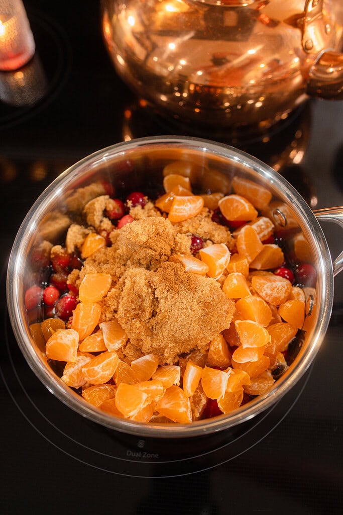 Overhead view of a saucepan on the stovetop filled with fresh cranberries, chopped mandarin orange segments, and brown sugar before cooking, with a glowing copper kettle and candle in the background.
