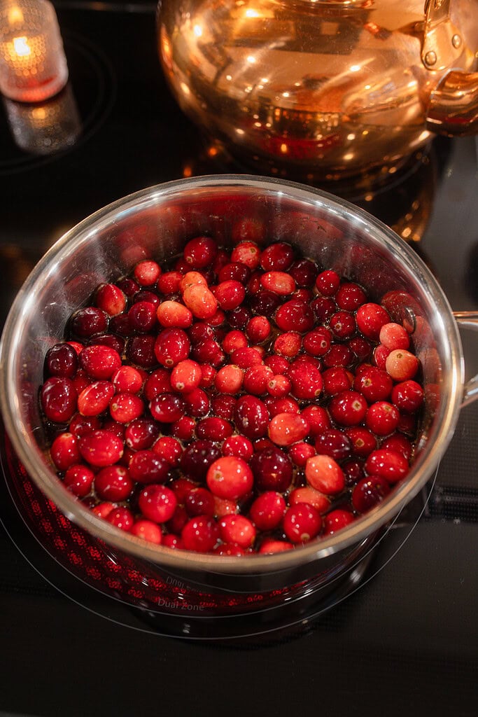 Overhead view of a saucepan filled with whole fresh cranberries simmering on the stovetop, with a glowing copper kettle and lit candle in the background.