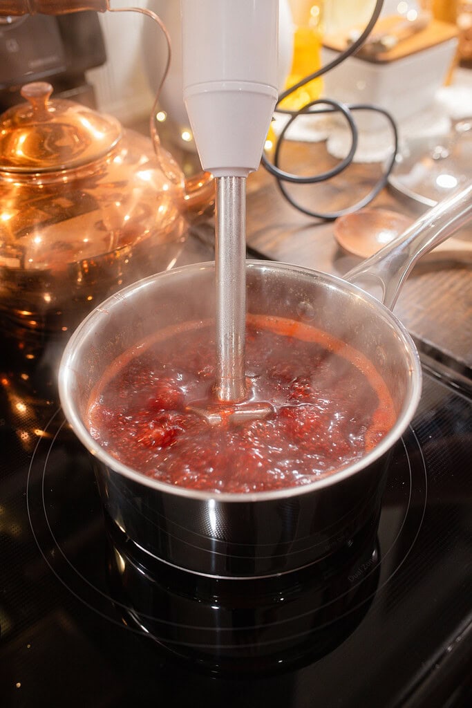An immersion blender partially blending a bubbling pot of cranberry orange sauce on the stovetop, with a copper kettle glowing in the background in a cottage kitchen.
