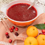 A vintage orange and white Pyrex dish of homemade cranberry orange sauce with a silver spoon, styled on a dusty rose linen with scattered fresh cranberries, mandarin oranges, a bay leaf, and dried baby's breath on a lace doily.