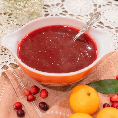 A vintage orange and white Pyrex dish of homemade cranberry orange sauce with a silver spoon, styled on a dusty rose linen with scattered fresh cranberries, mandarin oranges, a bay leaf, and dried baby's breath on a lace doily.