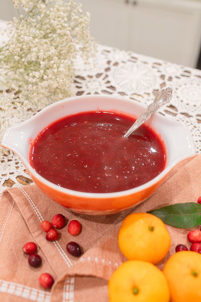 A vintage orange and white Pyrex dish of homemade cranberry orange sauce with a silver spoon, styled on a dusty rose linen with scattered fresh cranberries, mandarin oranges, a bay leaf, and dried baby's breath on a lace doily.