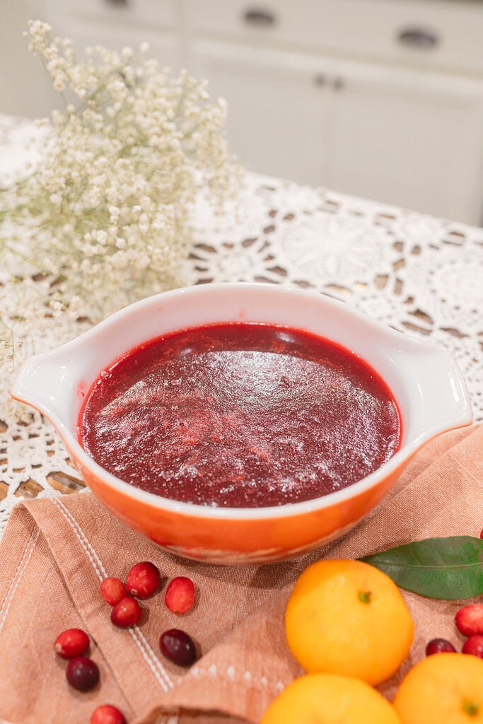 A vintage orange and white Pyrex dish filled with smooth homemade cranberry orange sauce, styled on a dusty rose linen with fresh cranberries, a mandarin orange, a bay leaf, and dried baby's breath in a cottage kitchen.