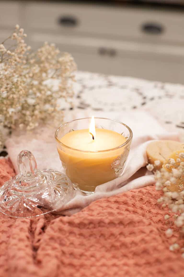 A glowing beeswax candle in a vintage glass jar, styled with its matching glass lid placed nearby. The setup includes a soft coral waffle-knit cloth, delicate baby’s breath flowers, and a cozy cottage-inspired background, creating a warm and serene atmosphere.