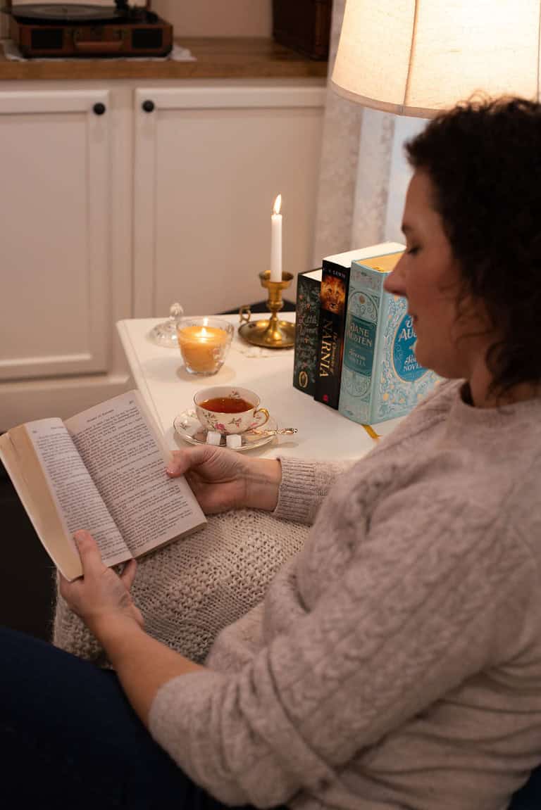 A woman reading Drums of Autumn by Diana Gabaldon while seated on a cozy armchair. A side table next to her features a teacup, sugar cubes, a lit candle, and a stack of books, with a warm brass candlestick completing the cozy setup.