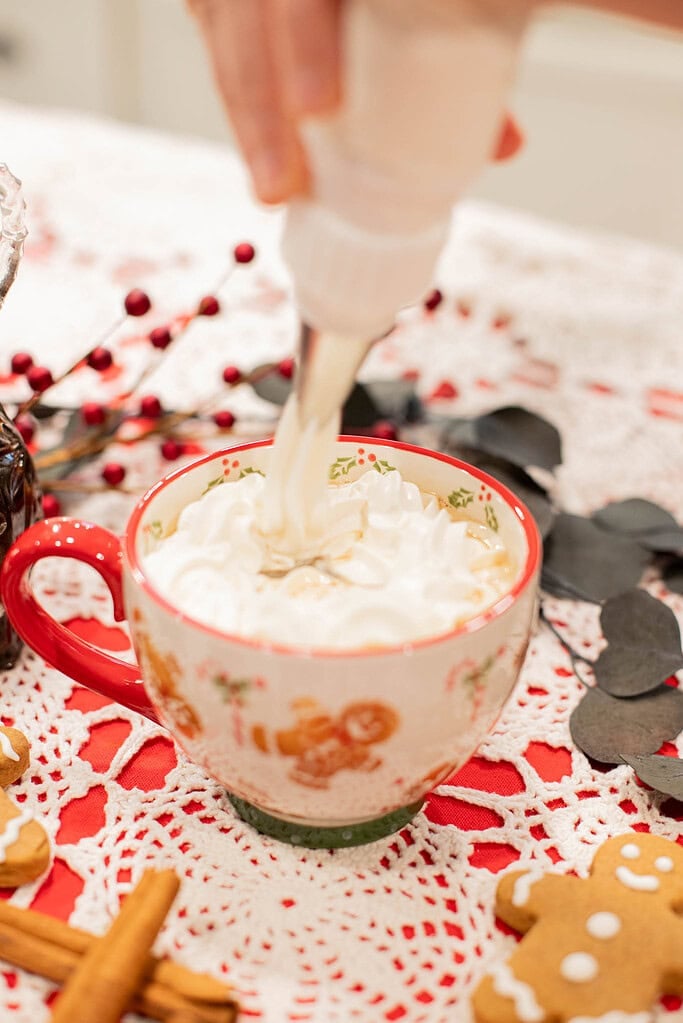 piping whipped cream onto gingerbread latte in festive mug