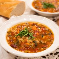 Two bowls of hearty minestrone soup served with a crusty baguette on the side, garnished with fresh basil and Parmesan cheese for a cozy presentation.