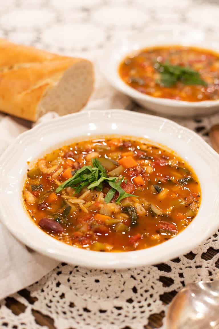Two bowls of hearty minestrone soup served with a crusty baguette on the side, garnished with fresh basil and Parmesan cheese for a cozy presentation.