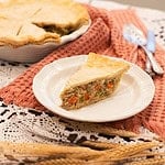 A full table view of a chicken pot pie served with a slice cut out, paired with a white plate and vintage utensils, on a charming lace tablecloth with rustic decor elements.
