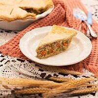 A full table view of a chicken pot pie served with a slice cut out, paired with a white plate and vintage utensils, on a charming lace tablecloth with rustic decor elements.