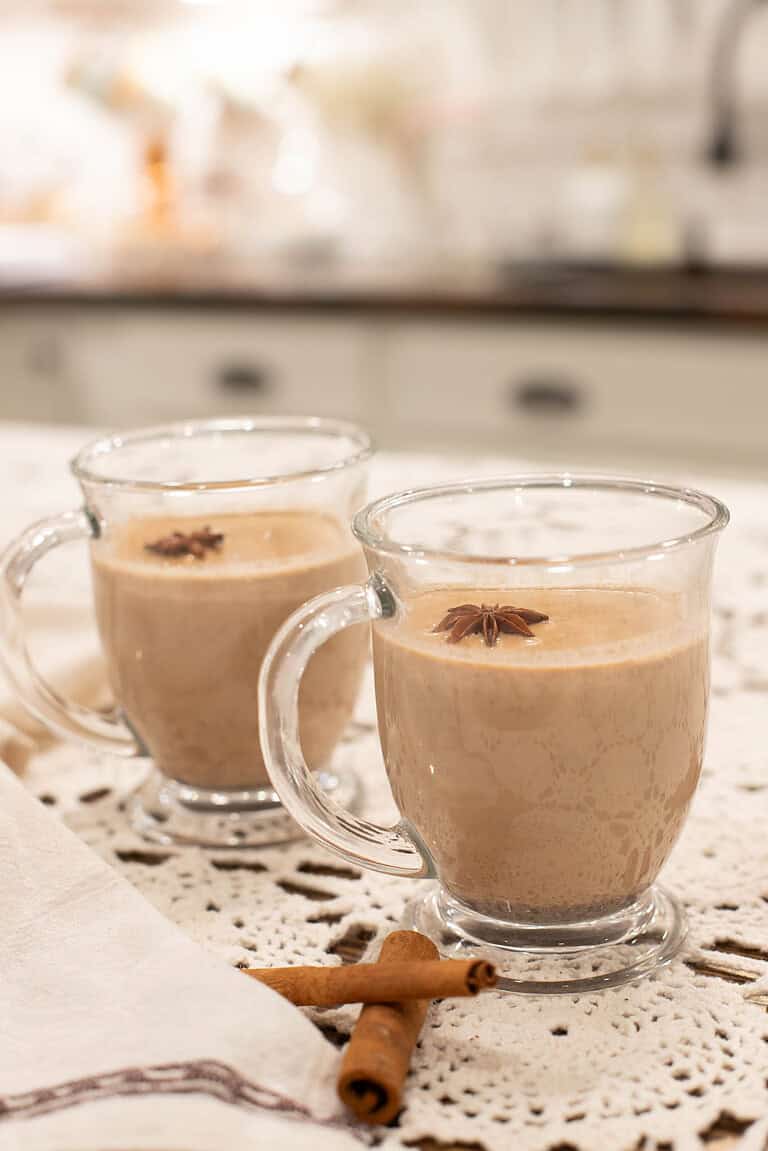 Two glass mugs filled with creamy chai latte, topped with star anise, set on a lace tablecloth for a cozy, inviting presentation.