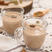 Another angle of two chai lattes, showing the frothy, spiced drink with star anise garnish, alongside scattered cinnamon sticks and cardamom pods for a rustic touch.