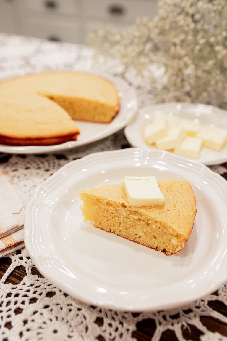 sourdough discard cornbread slice with butter on white plate with full cornbread in background