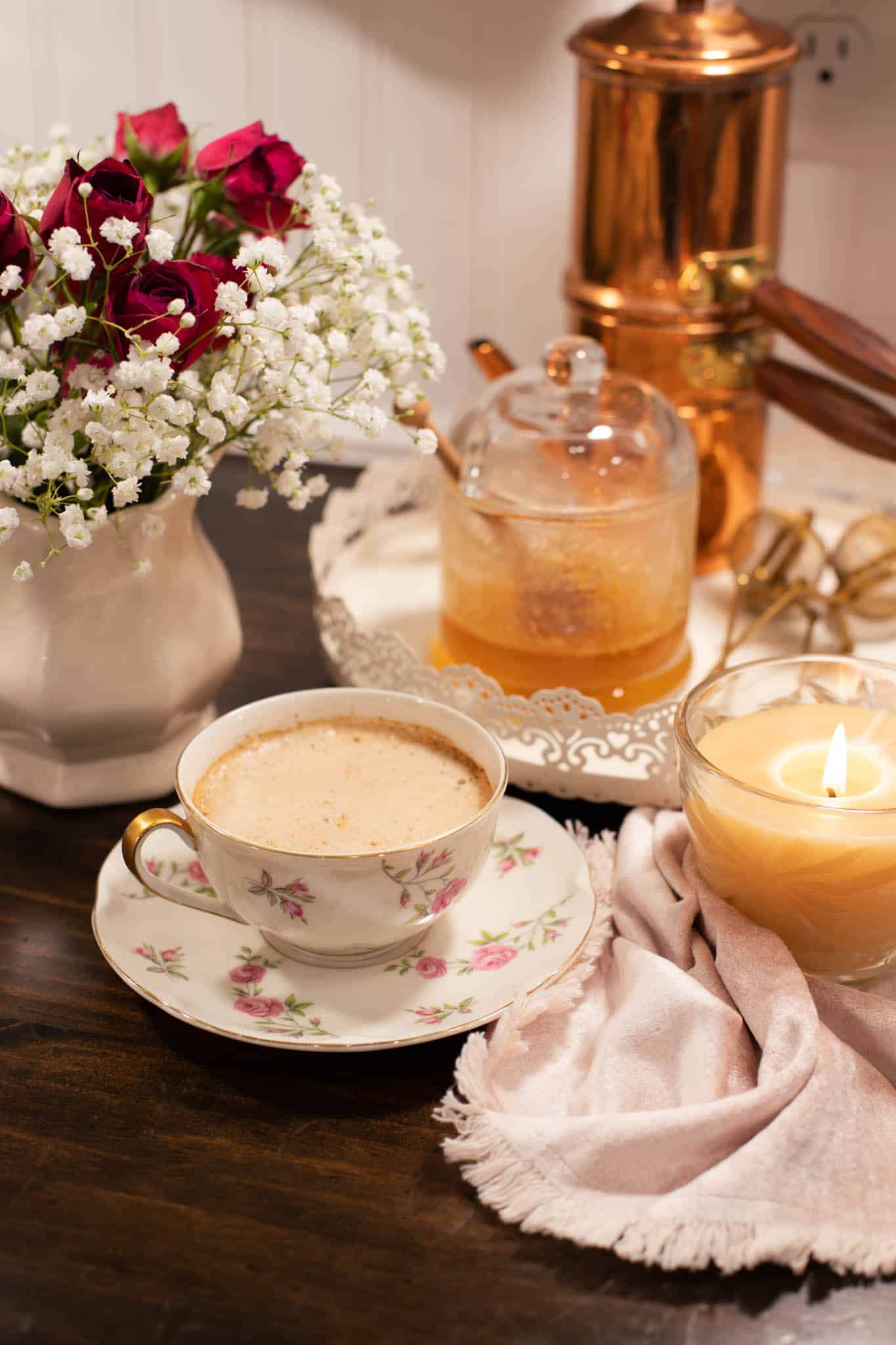 A cozy winter kitchen scene with a cup of frothy chai latte on a floral saucer, a lit candle, a honey jar, and a vase of red roses with baby’s breath on a rustic wooden table.