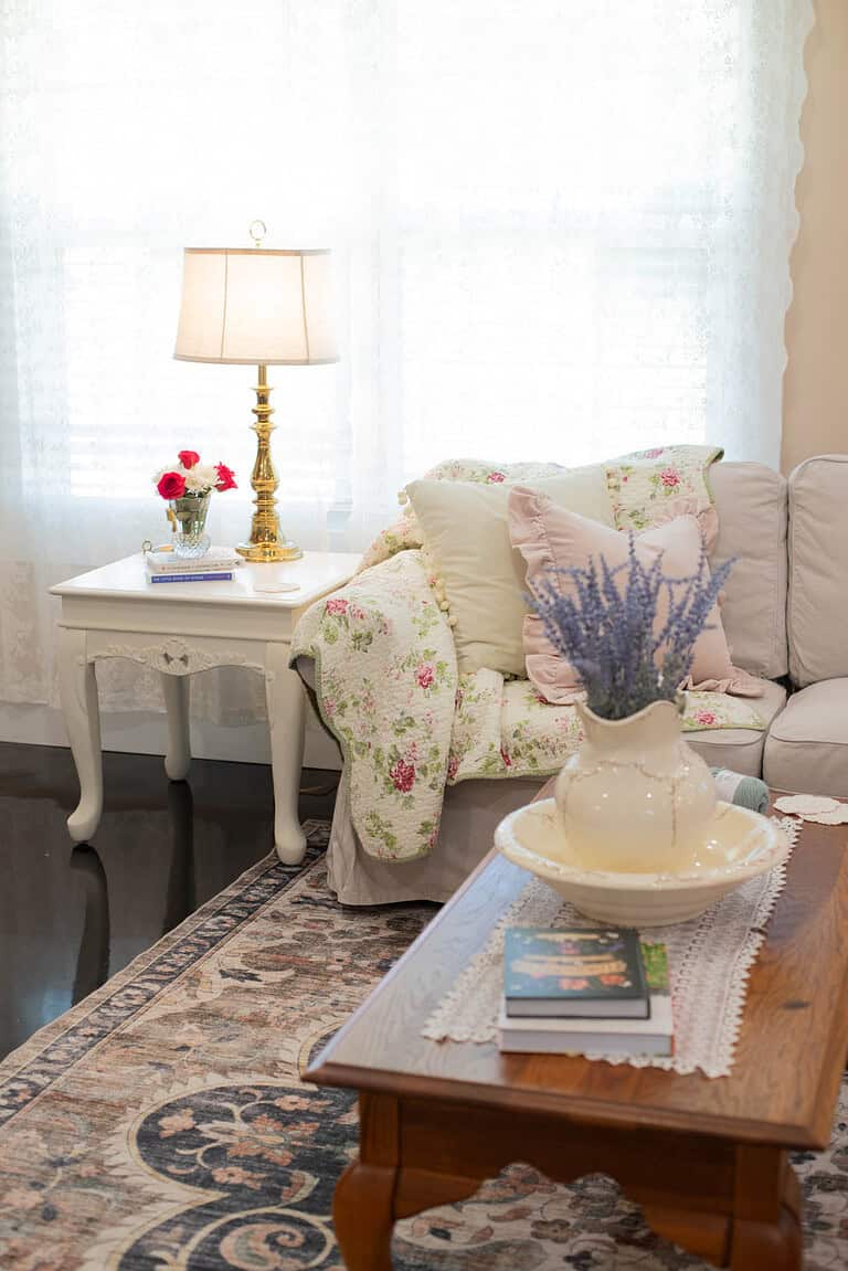 A cozy living room corner with a floral quilted throw, pastel pillows, and a vintage brass lamp on a lace-covered side table. A bouquet of lavender sits in a cream-colored pitcher on a wooden coffee table.
